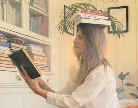 Picture of a the upper body of a white woman balancing a stack of books on her head while reading from another book in her hand.